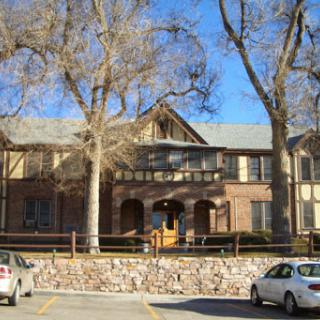 A charming two-story building surrounded by trees, showcasing a mix of architectural styles and a small parking area.