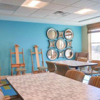 An art classroom with easels lined up against a blue wall, tables, and chairs arranged for students.