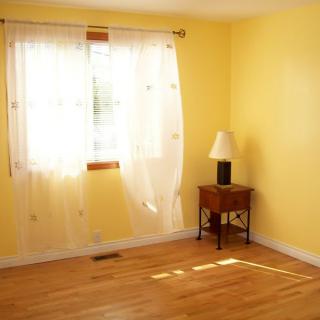 A bright, empty room with yellow walls and a wooden floor, featuring a window with white curtains and a small side table with a lamp.