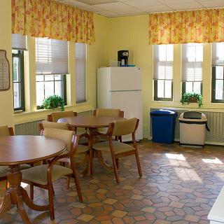 A cozy kitchen and dining area with round tables, chairs, large windows, and a refrigerator.