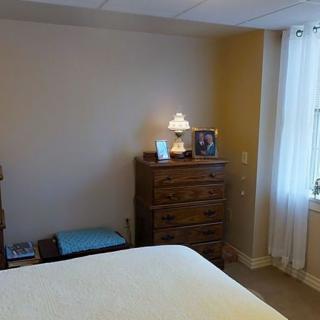 A cozy bedroom featuring a wooden dresser with a lamp and photo frame, alongside a window with sheer curtains.