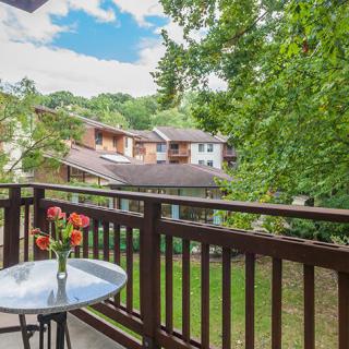 A cozy balcony with a small table and chairs, overlooking a view of trees and nearby buildings.