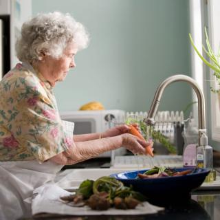 An elderly woman washes vegetables in the kitchen sink.