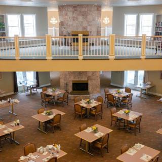 A spacious dining area with neatly arranged tables and chairs, featuring a central fireplace and an upstairs balcony.