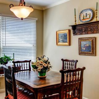 A cozy dining room with a wooden table and chairs, decorated with flowers and paintings, illuminated by natural light.
