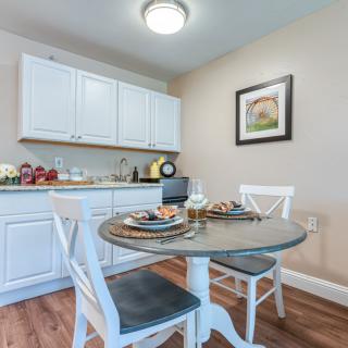 A cozy kitchen dining area featuring a small round table with two white chairs, surrounded by white cabinets and adorned with kitchen decor and artwork.