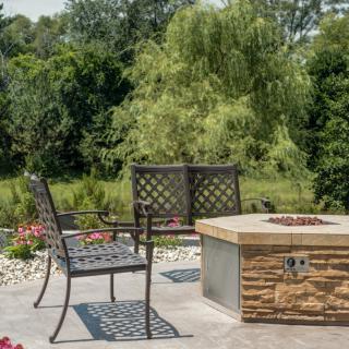 A serene patio setting with a stone fire pit and metal chairs, surrounded by lush greenery.