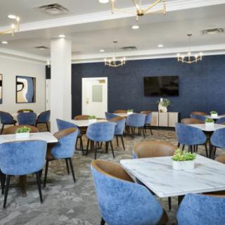 A modern dining area with blue chairs and white tables, featuring elegant chandeliers and a TV screen on the wall.