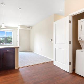 An open apartment kitchen with a stacked washer and dryer next to it, featuring wooden floors and ample natural light.
