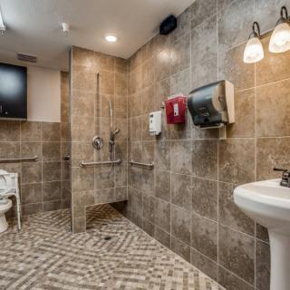 A modern, accessible bathroom with tiled walls and floor, featuring handrails, a sink, and dispensers.
