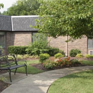 A peaceful garden area with a bench and pathway surrounded by greenery and a building in the background.