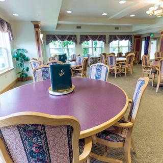 A well-lit dining area with multiple tables and chairs, featuring large windows and carpeted floors.