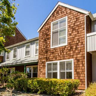 A two-story house with shingle siding and large windows, surrounded by trees and bushes.