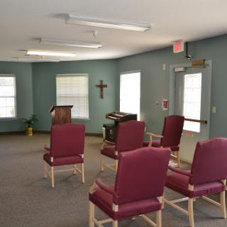A serene interior space featuring a few chairs arranged around a small podium, with a cross on the wall.