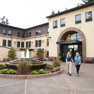 Two people walking near a building entrance with a fountain and landscaping.