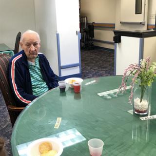 An elderly man sitting at a round table in a dining area, enjoying snacks and surrounded by a calm atmosphere.