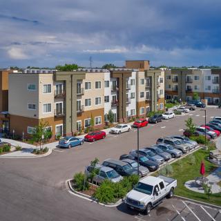 A modern apartment complex with a large parking lot and several parked cars under a cloudy sky.