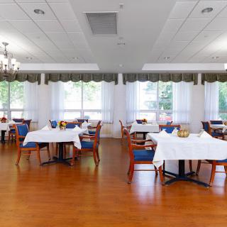 A bright and spacious dining room with elegantly set tables and chairs, featuring large windows and chandeliers.