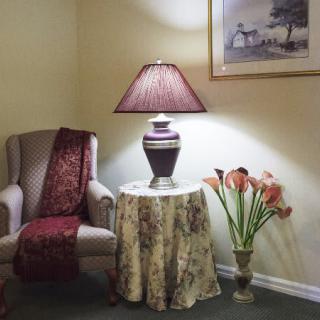 A cozy corner featuring an upholstered chair, a table with a decorative lamp, and a flower arrangement.