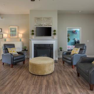 A cozy living room featuring gray armchairs, a central yellow ottoman, and a fireplace with a mounted television.