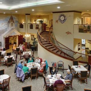 A spacious dining area with seniors gathered around tables, featuring a grand staircase and cozy ambiance.