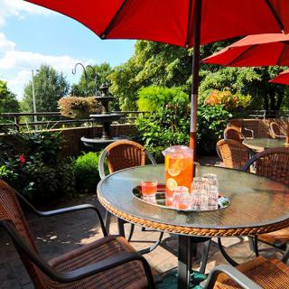 A sunny patio with a table set for refreshments under red umbrellas, surrounded by a lush garden and fountain.