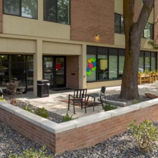 An outdoor seating area next to a brick building with large windows and a tree.
