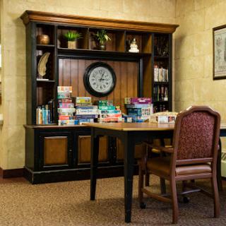 A cozy reading corner featuring a wooden shelf stocked with board games and a clock, accompanied by a table and chair.