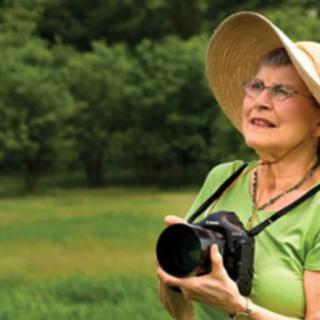 A woman wearing a wide-brimmed hat holds a camera outdoors in a grassy area.