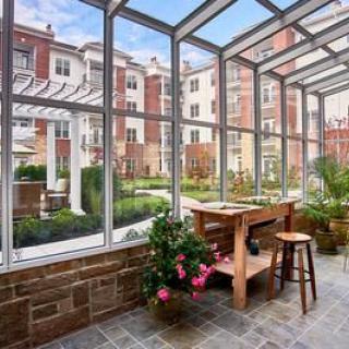A serene greenhouse space with wooden furniture and potted plants, overlooking a courtyard in an urban residential area.