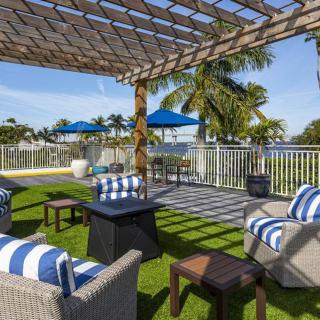 A stylish outdoor patio with comfortable seating under a pergola, surrounded by palm trees and blue skies.