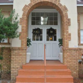 A brick building featuring an arched entrance with white double doors, flanked by decorative plants on steps.