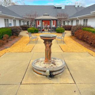A peaceful courtyard featuring a central fountain and surrounding benches, framed by landscaped paths and buildings.