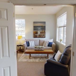 A cozy living room featuring a comfortable sofa and armchair, centered around a wooden coffee table. Large windows allow natural light to fill the space, while decorative pillows and wall art add a touch of style.