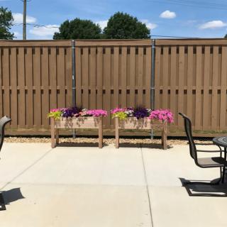 A sunny patio area with flower pots, chairs, and a table against a wooden fence.