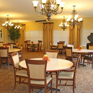 A well-lit dining area with round tables, comfortable chairs, chandeliers, and decorative plants.