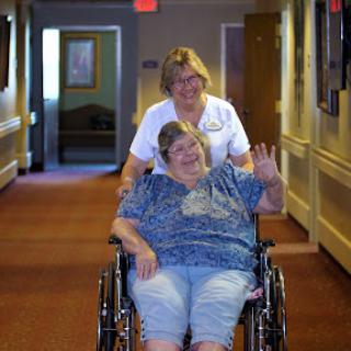 A caregiver helps an elderly woman in a wheelchair navigate the hallways of a healthcare facility.