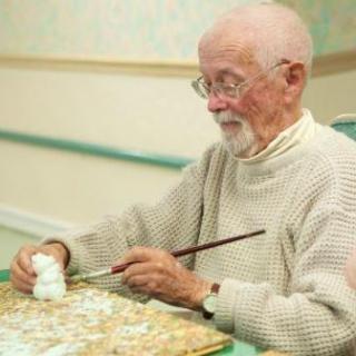 An elderly man deeply focused on painting a small object indoors.