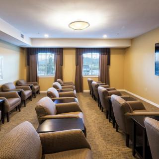 A small conference or meeting room with rows of comfortable chairs, natural light from two windows, and warm lighting.