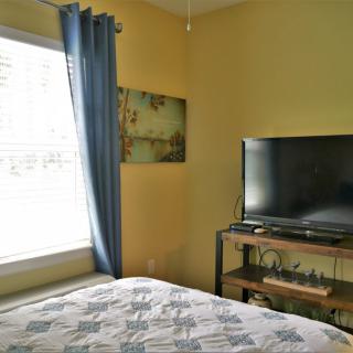 A cozy bedroom with yellow walls, featuring a bed, a TV on a stand, and a window adorned with blue curtains.