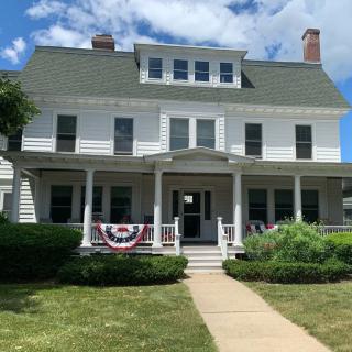 A large white house with a porch decorated with American flag bunting sits under a partly cloudy sky.