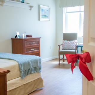 A cozy bedroom with a neatly made bed, wooden dresser, and a chair near the window. A red bow is tied to the door handle.