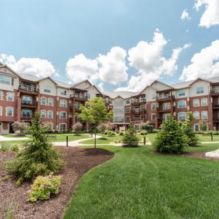 A picturesque apartment complex with lush greenery and a well-maintained lawn under a partly cloudy sky.