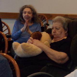 Two elderly women seated indoors, one holding a teddy bear.