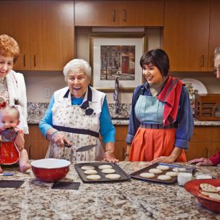 A group of people baking cookies together in a kitchen, all wearing aprons and smiling.