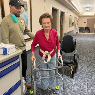 An elderly woman confidently using a walker with assistance in a healthcare setting.