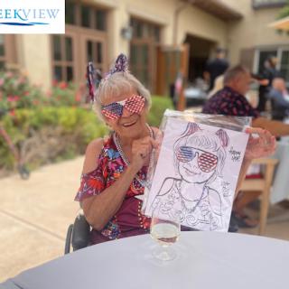An elderly woman proudly displays her colorful drawing while enjoying a sunny day outdoors.