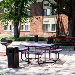 An outdoor picnic area with a table and benches next to a grill, situated in front of an apartment building on a sunny day.