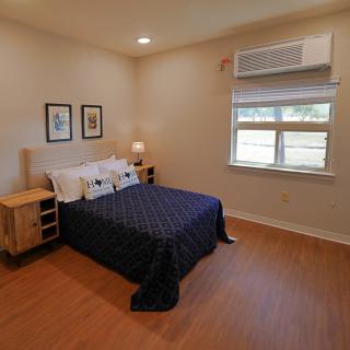 A minimalist bedroom with a neatly made bed, wooden flooring, nightstands, and wall art. The room is well-lit with natural light coming through the window.