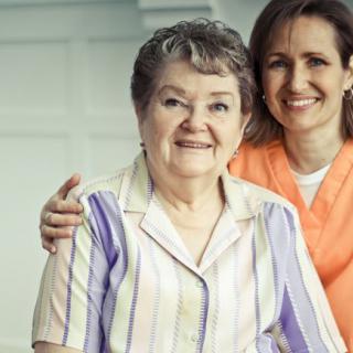 An elderly woman smiling with her caregiver by her side.